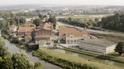 Luftaufnahme einer Fabrikanlage mit roten Backsteingebäuden und roten Dächern, gelegen an einem Fluss, der in der linken unteren Bildecke zu sehen ist. Im Hintergrund erstreckt sich eine Stadt mit Wohngebieten und landwirtschaftlichen Flächen.