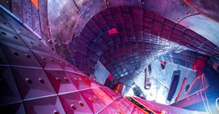Blick in das gewundene Plasmagefäß des Stellarators Wendelstein 7-Xin rötlichem und blauem Licht. Die Wände sind mit rechteckigen Kacheln ausgekleidet.